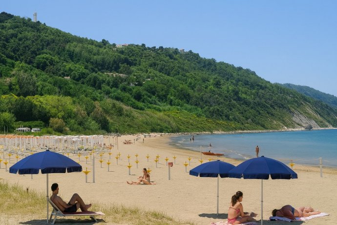 Spiaggia Baia Flaminia Pesaro con ombrelloni blu e bagnanti sul litorale sabbioso.