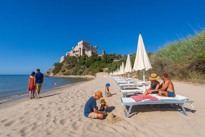 Spiaggia di Falconara con Castello, ombrelloni bianchi e bagnanti su sabbia dorata.