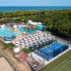Complesso turistico con piscine, scivoli, ombrelloni, campi sportivi e vista sul mare in lontananza.