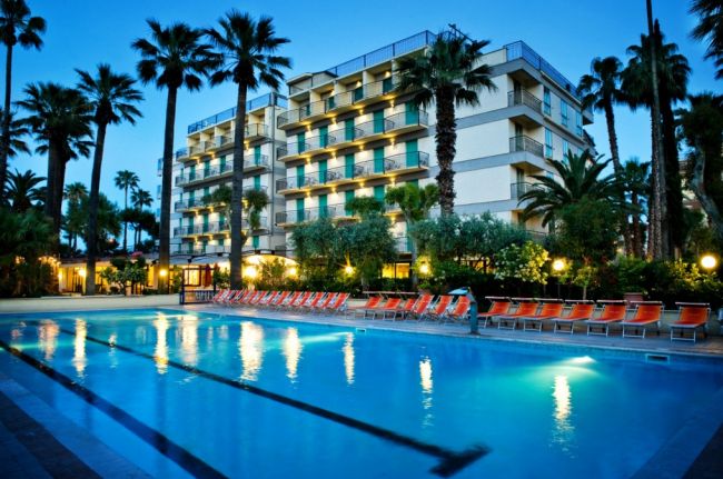 Hotel con piscina illuminata di sera, circondato da palme e lettini arancioni.