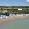 Spiaggia con ombrelloni blu, bagnanti, mare calmo, colline verdi sullo sfondo.