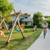 Bambini su altalene in un giardino, persona in bici su vialetto di ghiaia.