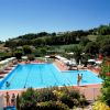 Piscina con ombrelloni, persone a bordo e colline verdi attorno.