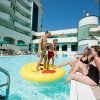 Due ragazzi su un materassino in piscina, due donne sul bordo, palazzi sullo sfondo.