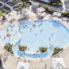 Piscina affollata con bambini che giocano, circondata da ombrelloni bianchi e area verde.