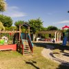 Un parco giochi per bambini con scivolo, altalena e aree per sedersi, circondato da alberi.